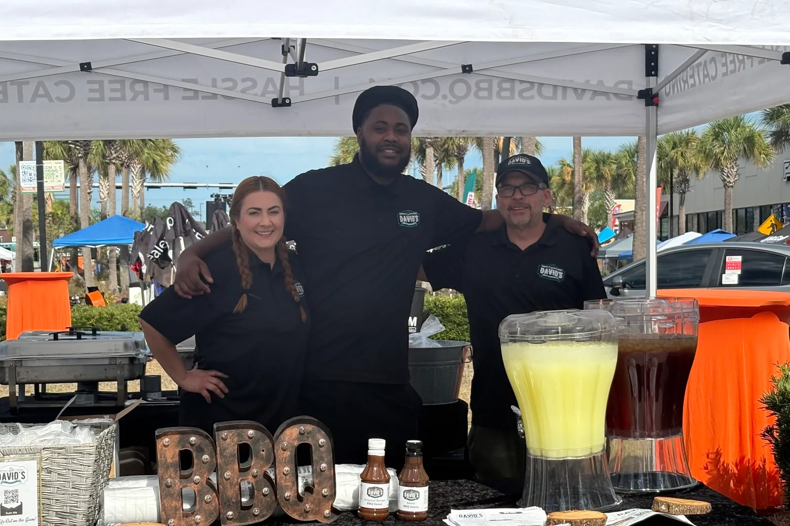 Three of the catering crew standing behind a table of sauce bottles and drink dispensers under a tent, beside metal letters spelling BBQ.