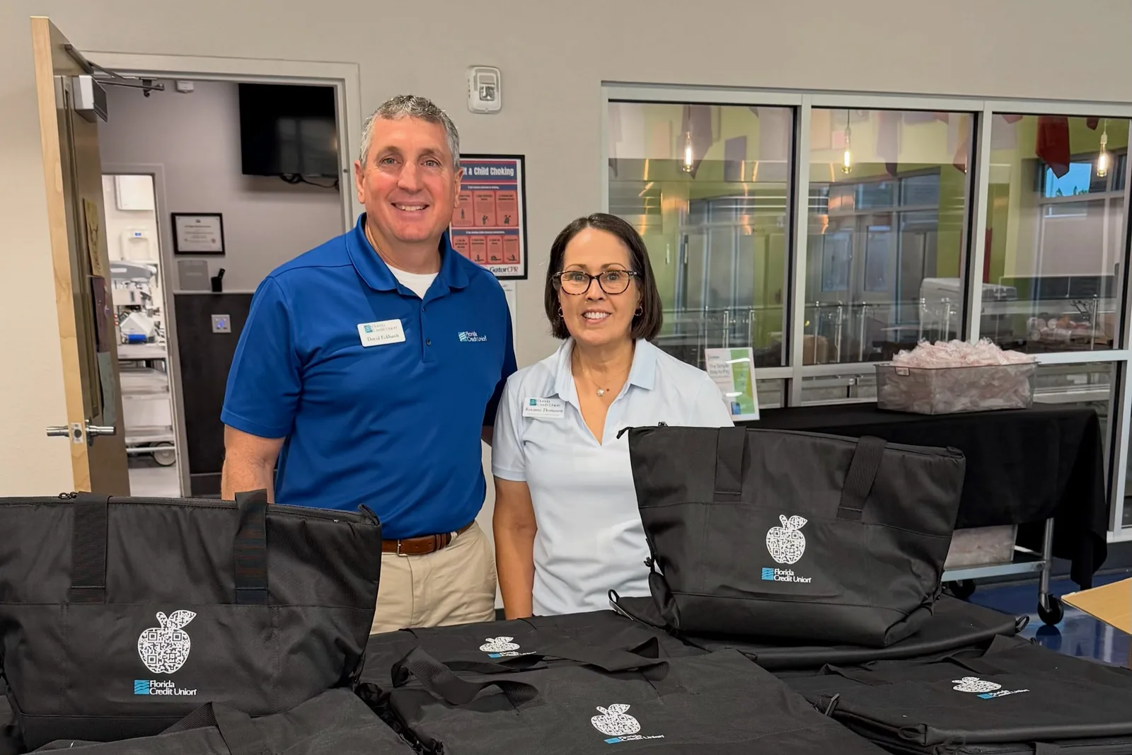 Two people standing behind a table stacked with insulated catering bags in an office lobby.