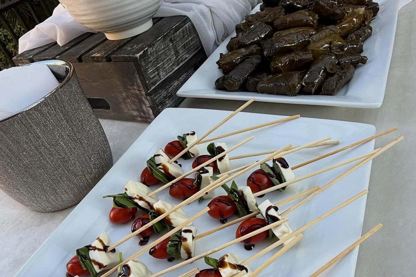 Caprese skewers laid out on a white platter beside a tray of stuffed grape leaves and bowls of cut fruit.