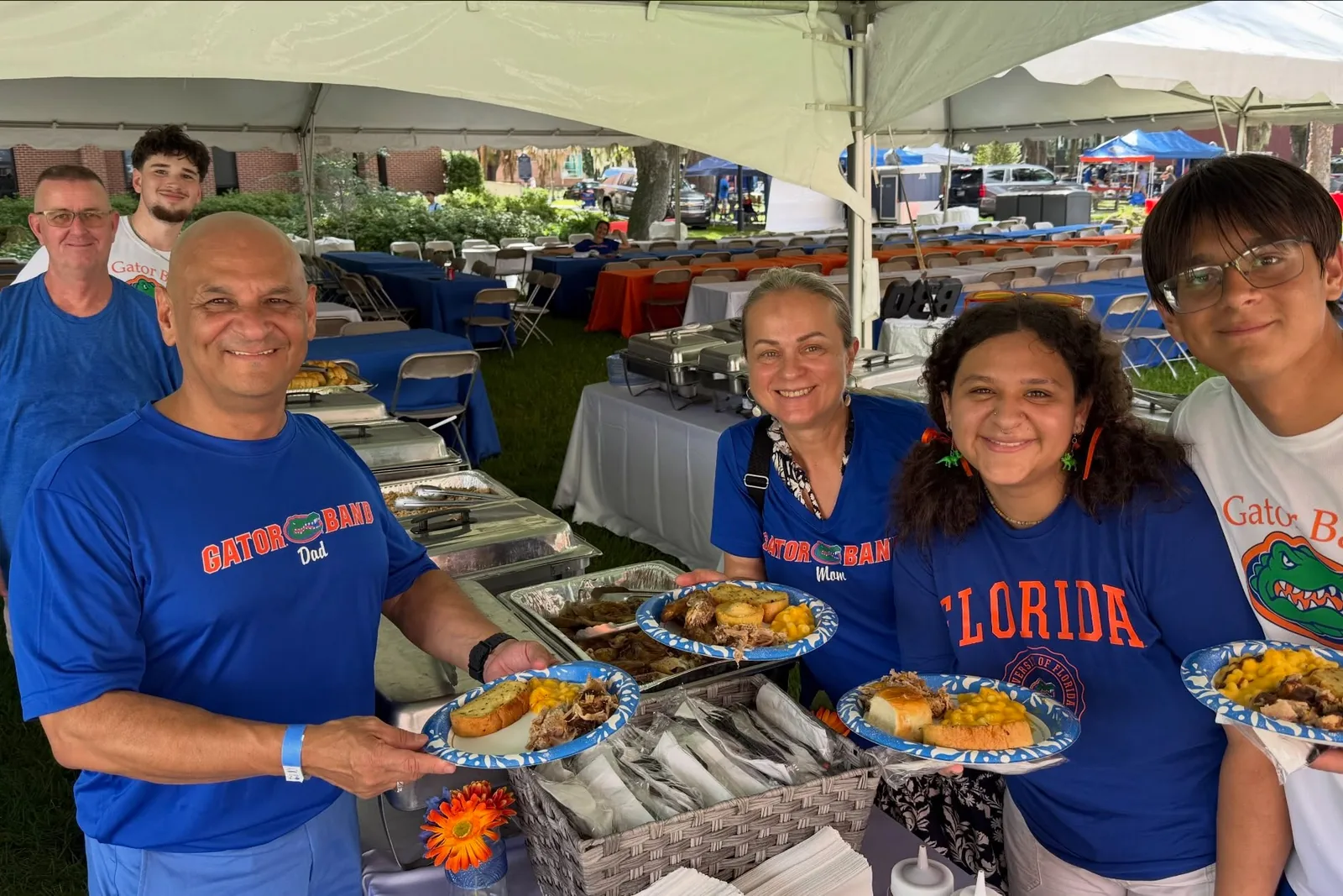 Six people holding plates of barbecue at a tailgate under a white tent, with chafing dishes on the table behind them.