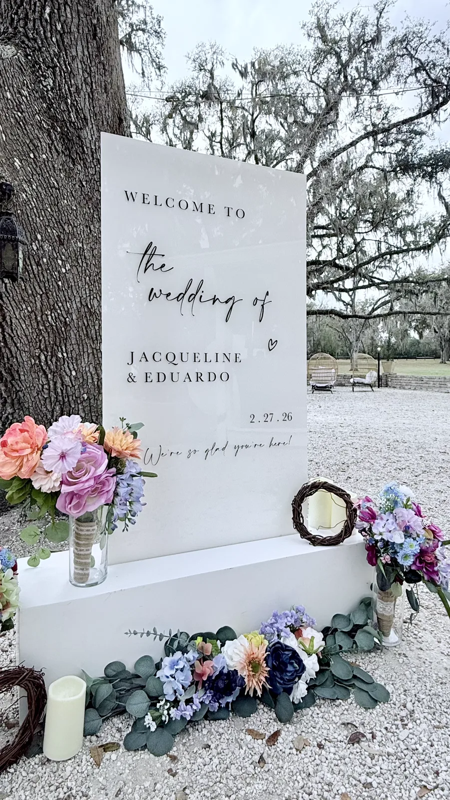 A white welcome sign on a pedestal with bright florals beneath a live oak