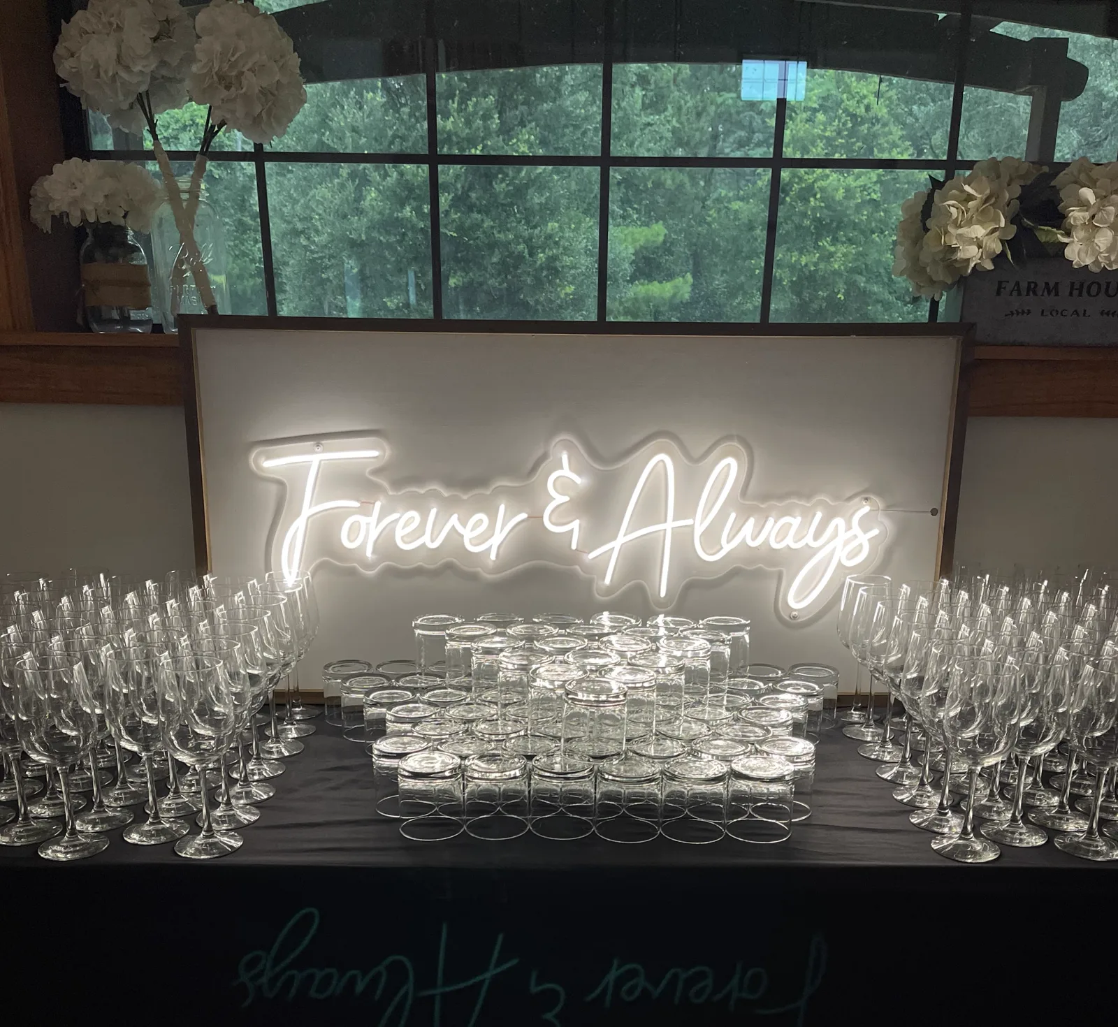 A Forever and Always neon sign above rows of wine glasses and stacked bar glasses