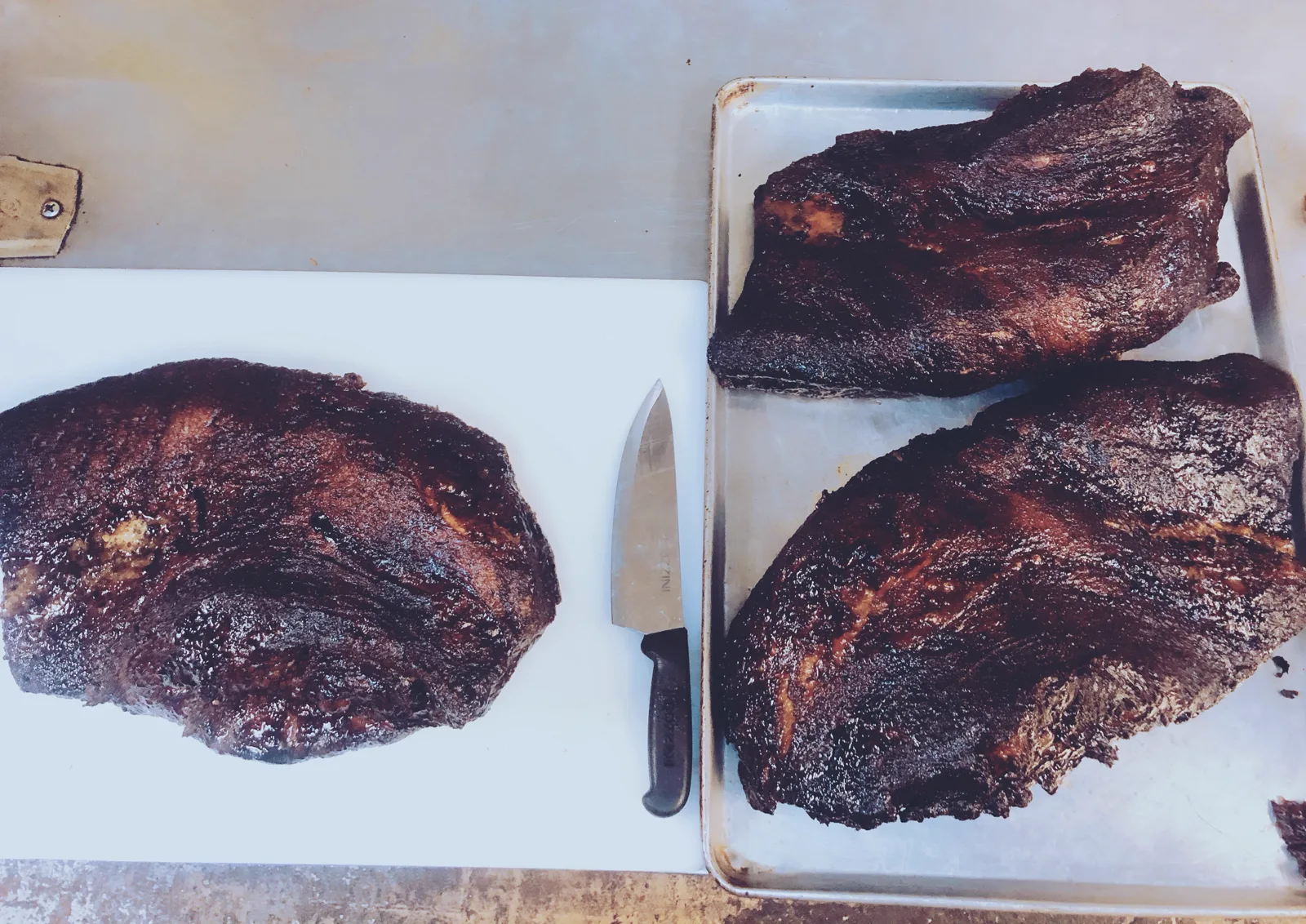 Three whole smoked briskets on a cutting board and sheet pan beside a chef's knife