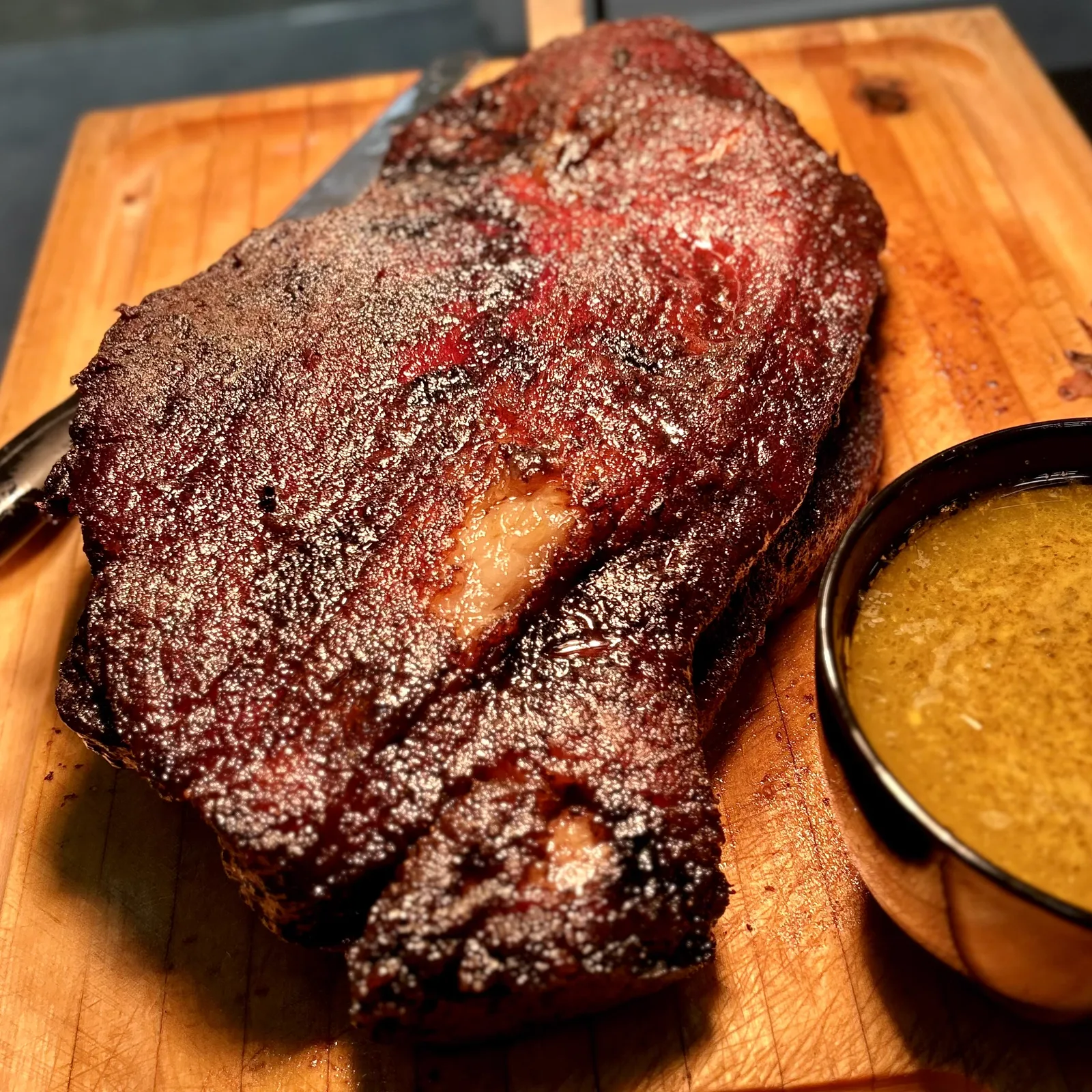 A peppered smoked roast on a carving board beside a bowl of golden sauce