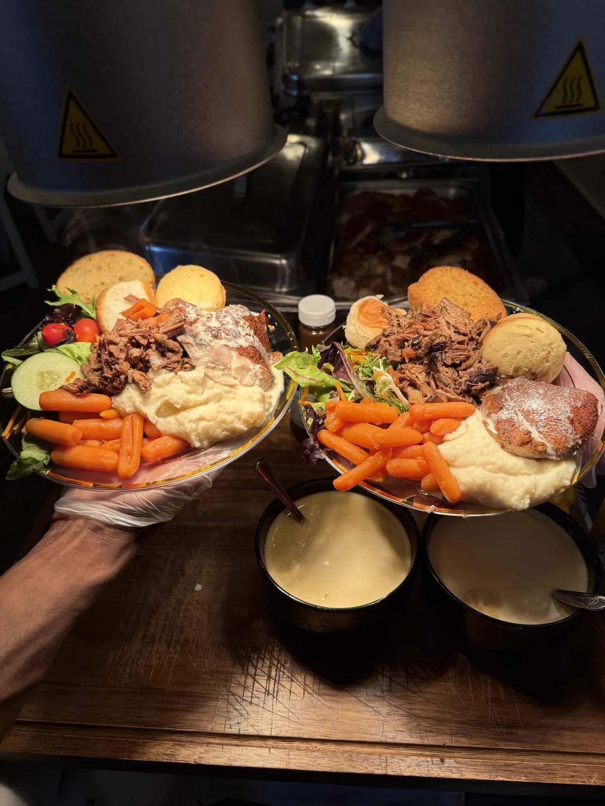 Two dinner plates of pulled smoked meat, mashed potatoes, carrots, salad, and rolls held above bowls of sauce