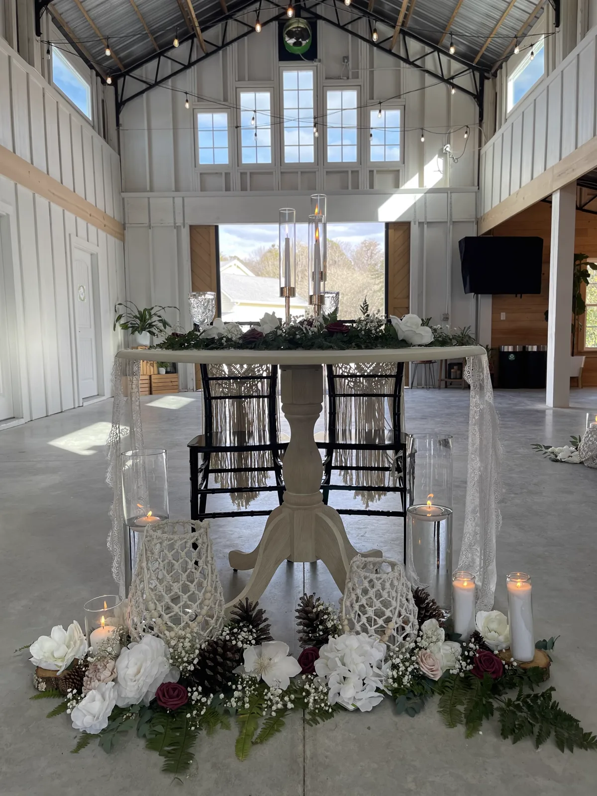 A round sweetheart table trimmed in lace with candles and florals in a white barn hall