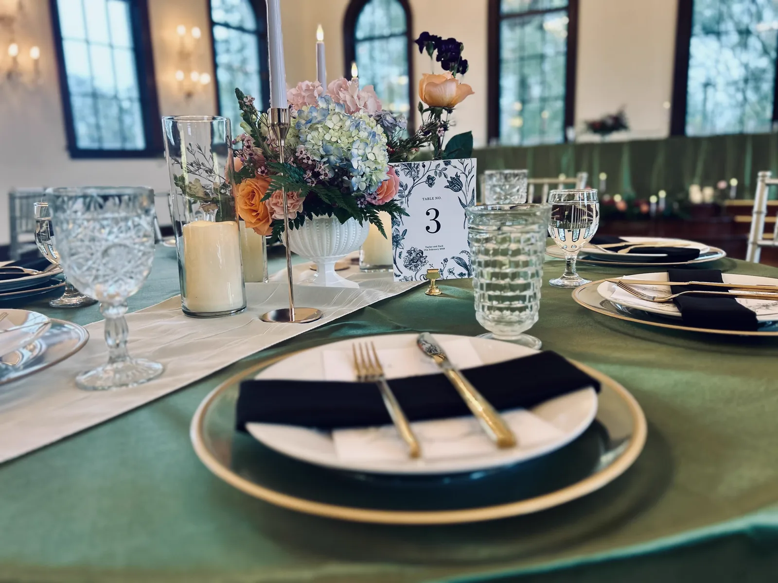 A sage green table with black napkins on gold rimmed plates, gold cutlery and pastel florals