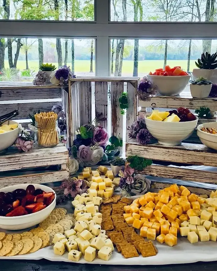 A grazing table on whitewashed crates by a wall of windows, bowls of watermelon, pineapple, grapes and strawberries above a long fan of cubed cheese, crackers and flowers