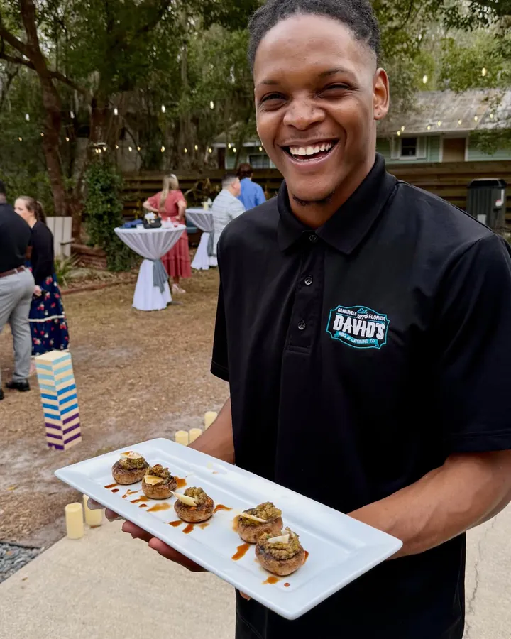 A server in a David's BBQ and Catering shirt smiling behind a long white platter of stuffed mushrooms at an outdoor event, string lights and cocktail tables behind