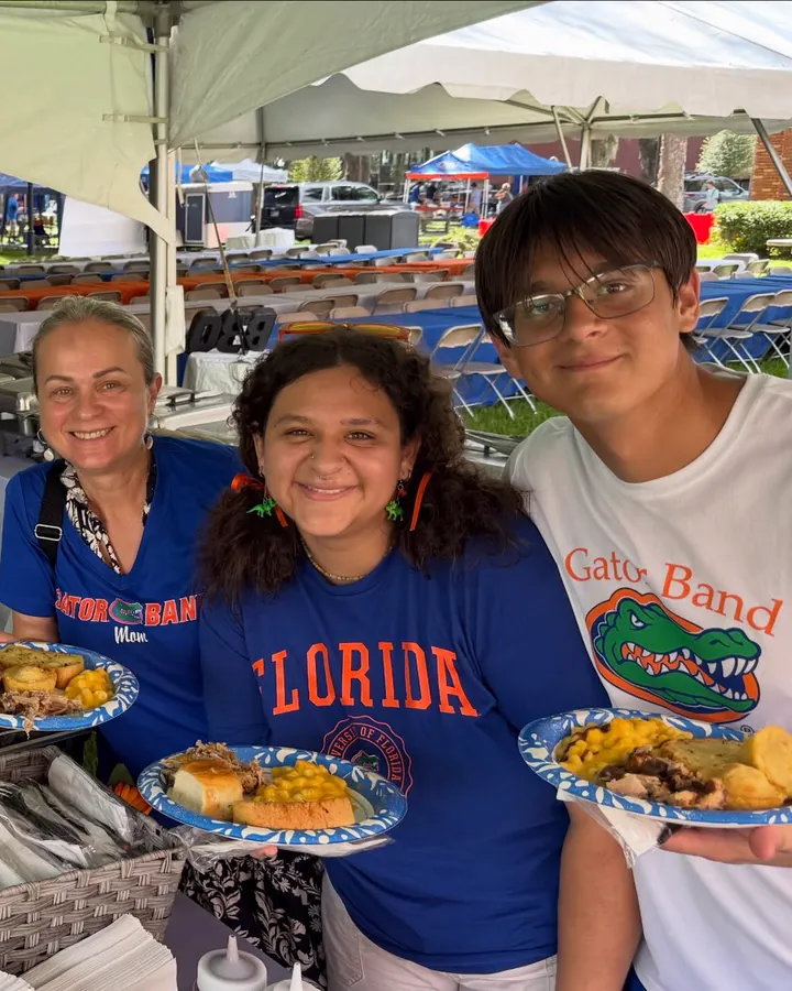 Three guests in Gators shirts holding plates of pulled pork, cornbread and macaroni under a catering tent, with chafing dishes on the table beside them