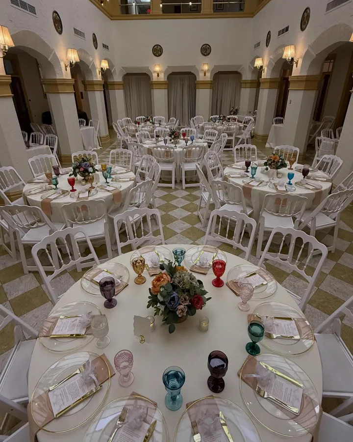 A ballroom set for a reception, round tables in ivory linen with gold rimmed chargers, colored glassware and low floral centerpieces, white chairs all around
