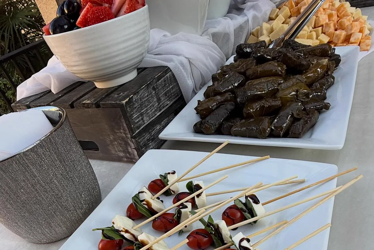 Caprese skewers, stuffed grape leaves, cubed cheese and melon, and a bowl of berries on an outdoor table.
