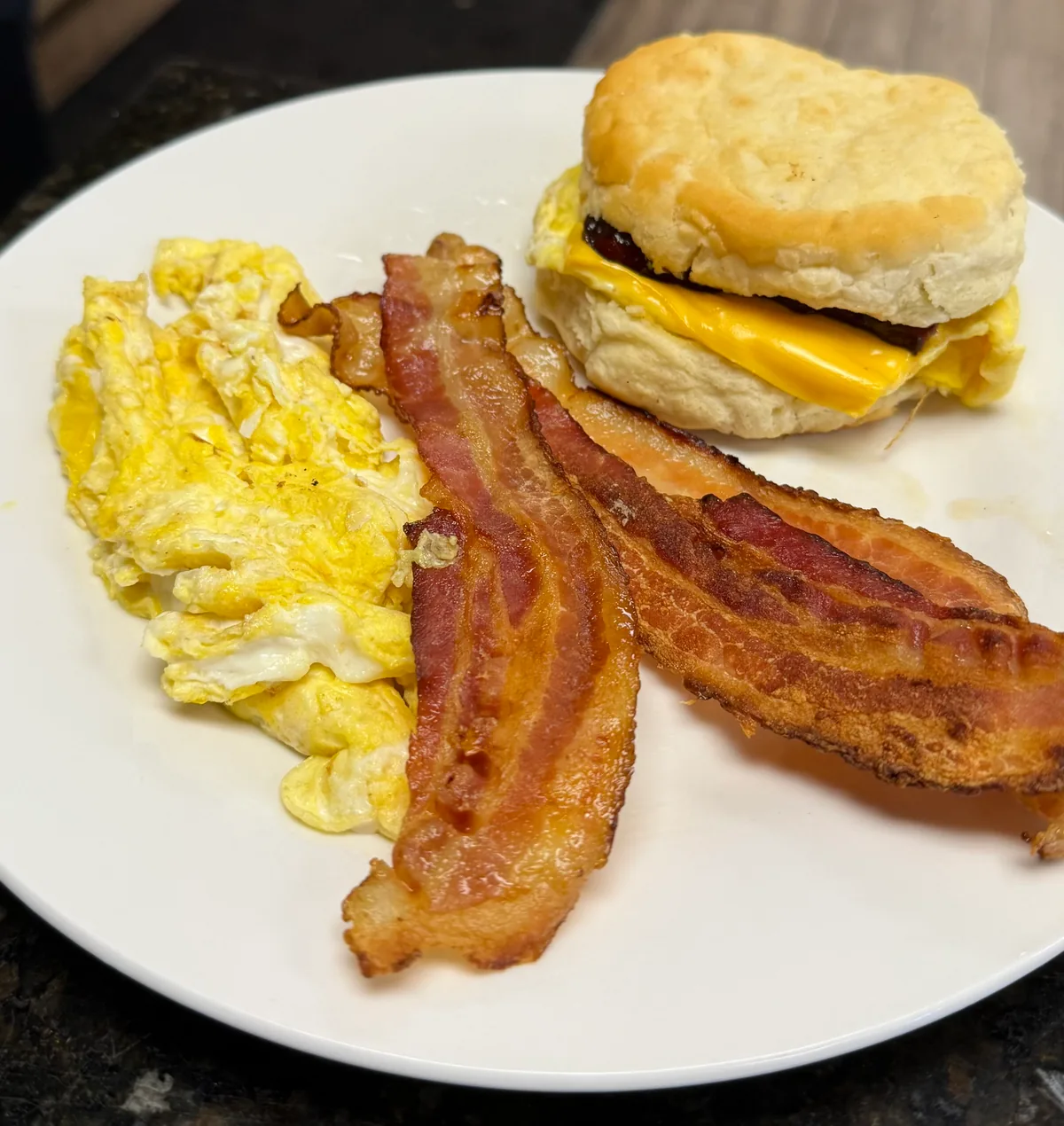 A breakfast biscuit with egg and cheese beside scrambled eggs and two strips of bacon on a white plate
