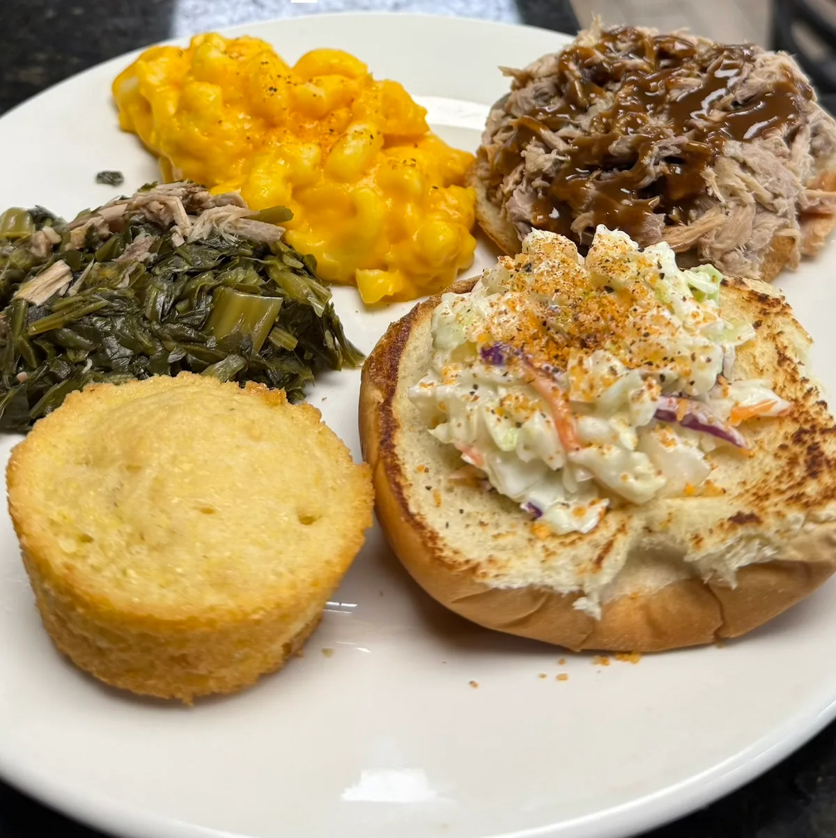 A pulled pork sandwich plate with slaw on the bun, mac and cheese, collard greens, and a cornbread muffin