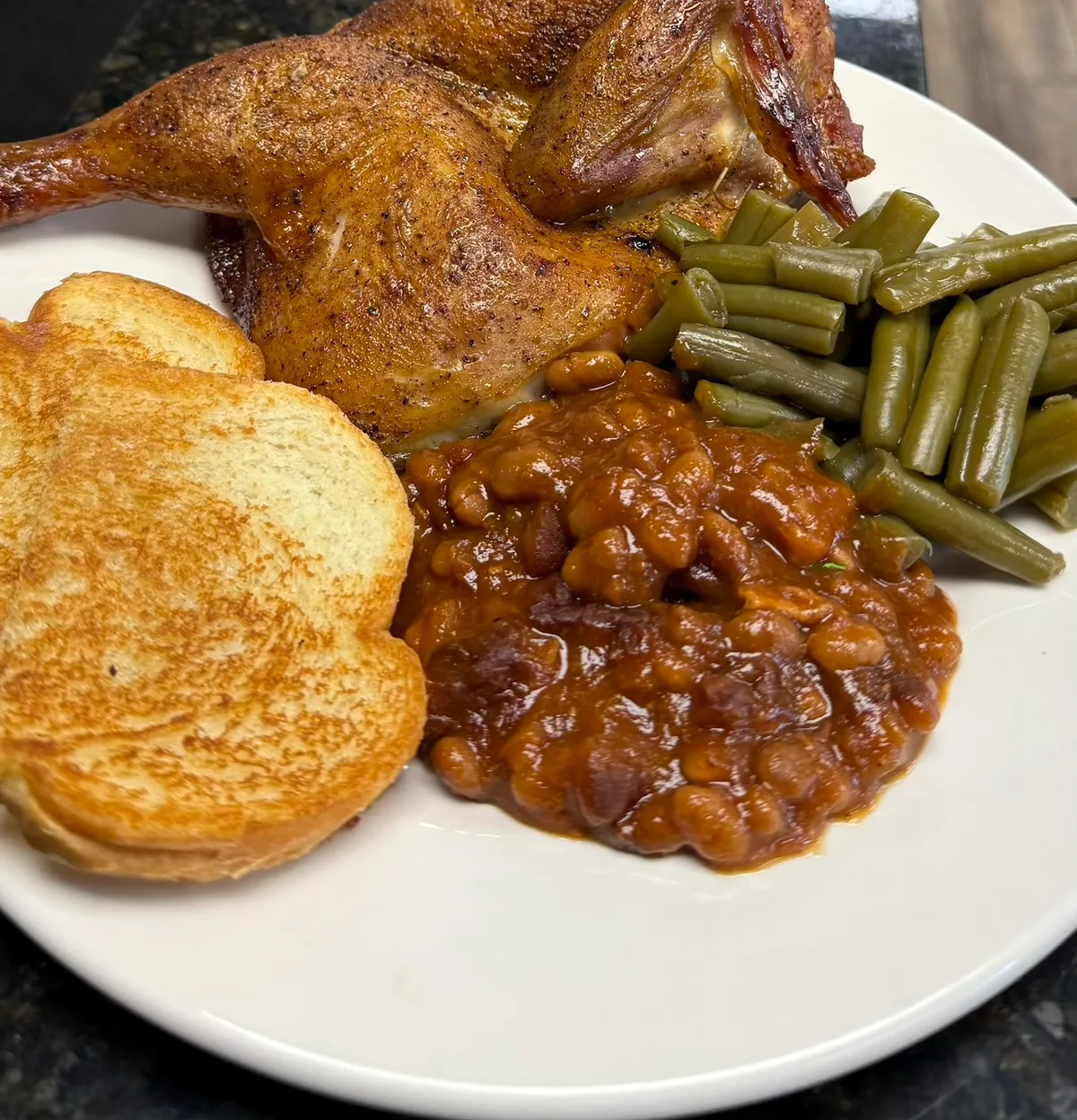 A smoked quarter chicken on a plate with baked beans, green beans, and Texas toast